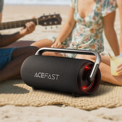 Black portable speaker with red accents on a sandy surface, people playing guitar in the background